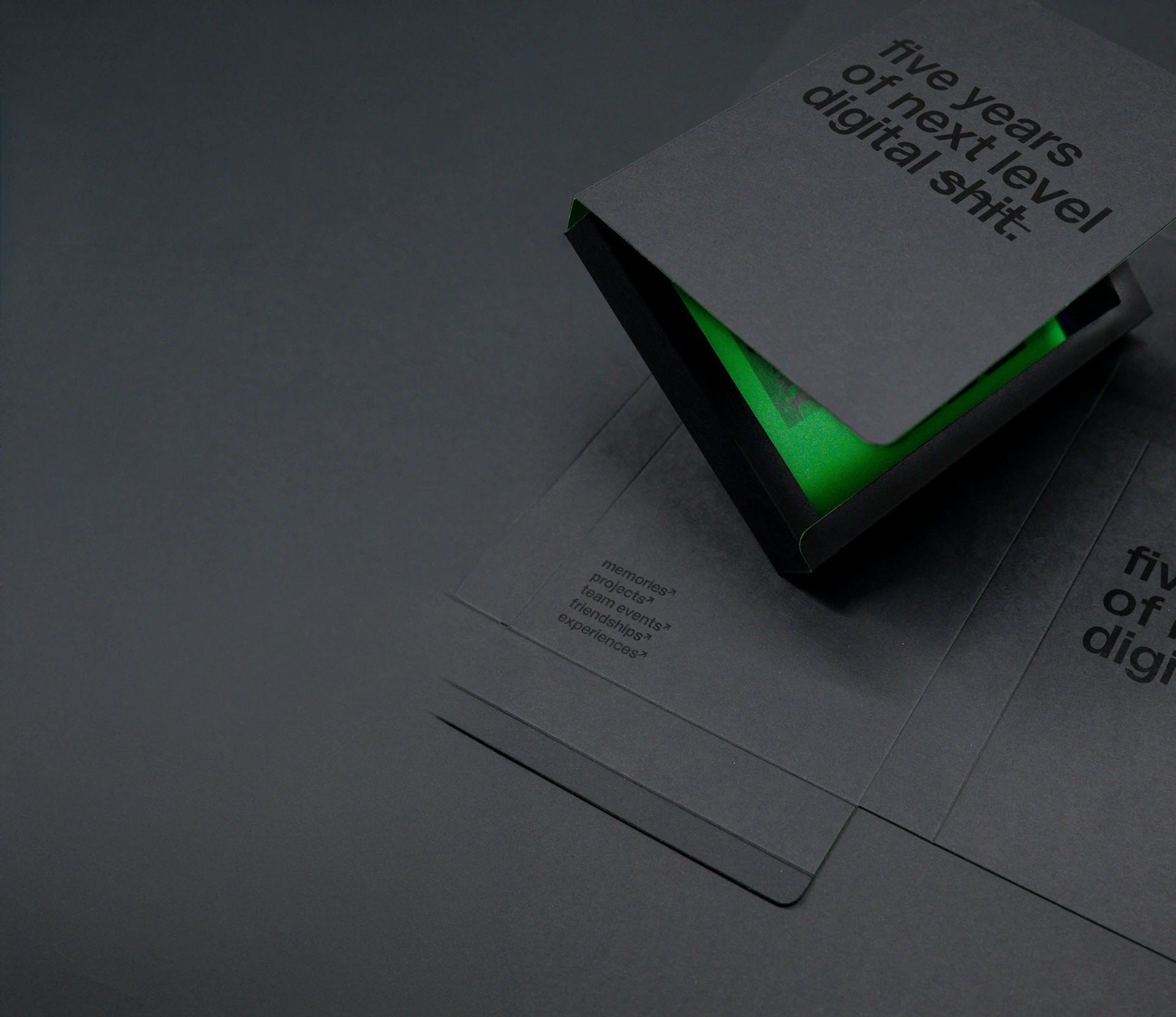 Schwarzes, teilweise geöffnetes Heft mit dem Text „five years of next level digital sh!t.” und grüner Innenseite, auf einer schwarzen Fläche liegend, darunter ein weiteres ähnliches Heft.