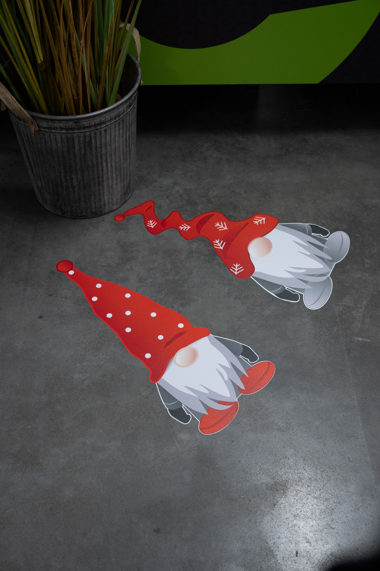 Zwei Cartoon-Zwerg-Aufkleber mit roten Hüten und weißen Bärten auf einem glatten grauen Boden neben einem Metalltopf mit hohen grünen Pflanzen.